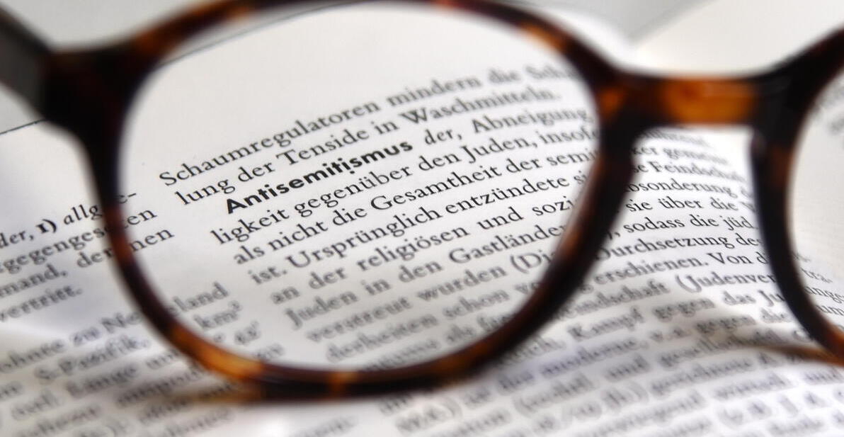 Das Bild zeigt eine Brille auf einem Buch, wo das Wort Antisemitismus hervorsticht