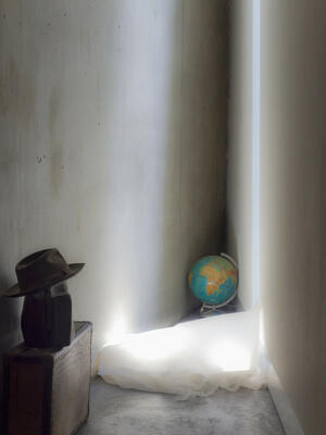 Bild vergrößern: Das Bild zeigt eine Zimmerecke,auf einem Koffer stehen eine Tasche und ein Hut, ein weißes Stück Stoff und ein Globus liegen auf dem Boden.