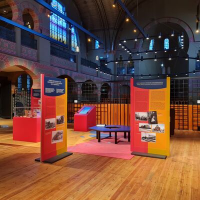 Bild vergrößern: Eine Ausstellungsvitrine, Ausstellungswände, ein bedruckter Ausstellungsteppich und ein Touchscreen sind zu sehen. Im Hintergrund ist die Synagoge in Groningen zu sehen.